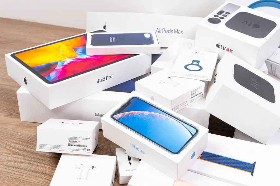 A pile of Apple product boxes, including iPhone, iPad Pro, AirPods, Apple Watch, and Apple TV, arranged on a wooden surface.